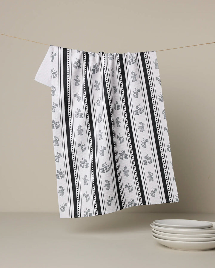 The Geometry Bunny Dots Stripe tea towel, featuring black vertical stripes and small floral patterns on white, hangs above a light surface next to four white plates—an ultra-absorbent addition to any modern kitchen.