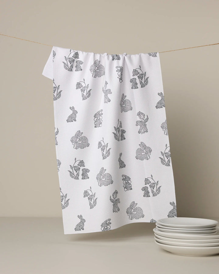The Geometry Bunny Dots tea towel, featuring quick-drying white fabric with black line drawings of rabbits and flowers, hangs beside a stack of white plates against a beige background.
