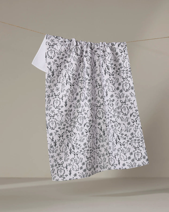 The Bunny Rabbit Garden towel by Charlie Rowan Designs hangs from a clothesline, its charming black floral pattern standing out against a simple, neutral background.