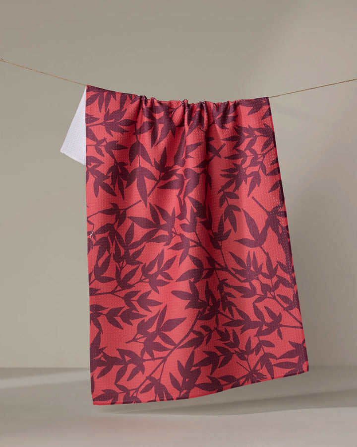 The Burgundy Branches tea towel by Rebecca Bobko, featuring a dark leaf pattern, hangs from a string against a beige background with one corner folded, showing its white underside and ultra-absorbent quality.