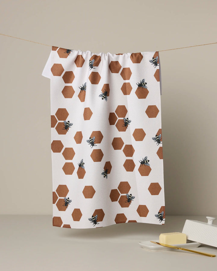 The Geometry Busy Buzzin tea towel, featuring a pattern of brown hexagons and bees, hangs on a line. In front, a stick of butter and covered butter dish sit on a beige surface.
