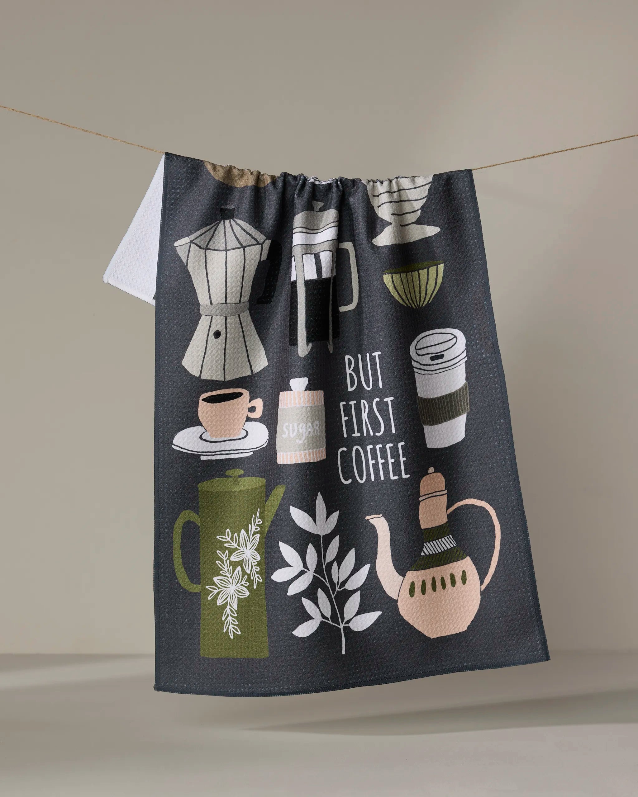 The "But First, Coffee" tea towel by Cottonflower Studio is made from recycled materials and features illustrations of coffee cups, a coffee pot, a sugar jar, and a French press, with the phrase “But First Coffee” displayed as it hangs.