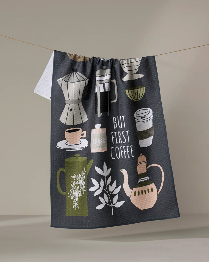 The "But First, Coffee" tea towel by Cottonflower Studio is made from recycled materials and features illustrations of coffee cups, a coffee pot, a sugar jar, and a French press, with the phrase “But First Coffee” displayed as it hangs.