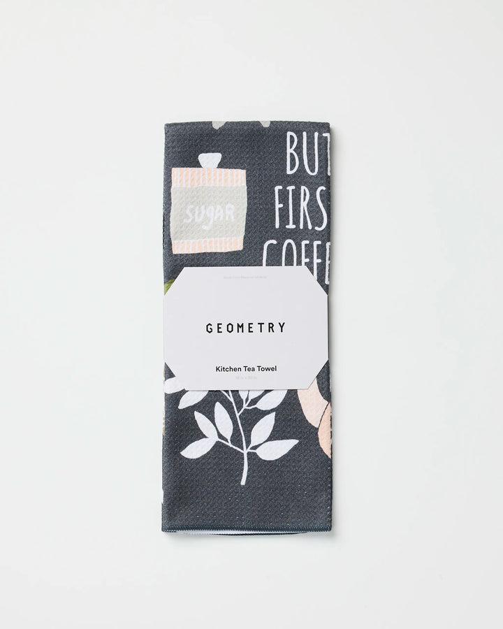 A folded tea towel made from recycled materials featuring a plant and text design, wrapped in a white label that reads "But First, Coffee" by Cottonflower Studio, placed on a plain white background.