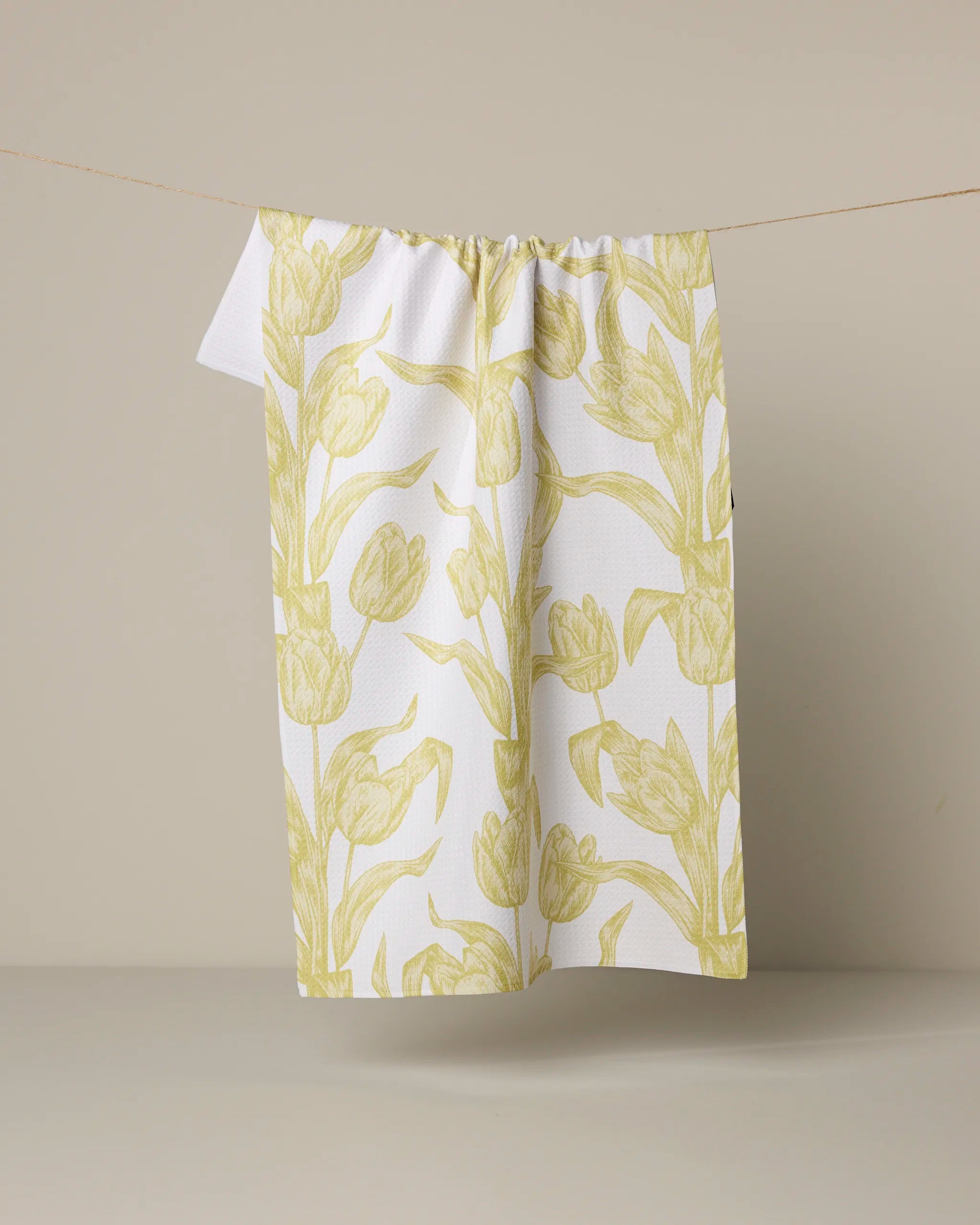 The Geometry "Butter Tulips" tea towel, featuring a white, ultra-absorbent fabric with a yellow tulip pattern, hangs on a line against a light beige background.