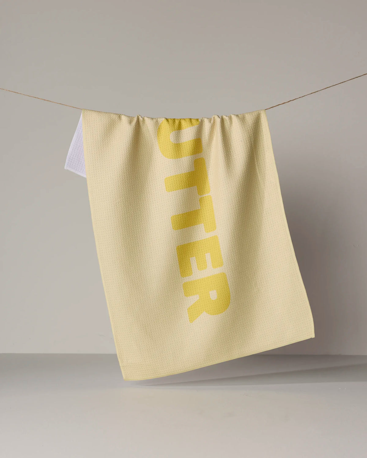A pale yellow, quick-drying GEOMETRY Butter tea towel hangs on a string, featuring the word BUTTER in large, bold letters.