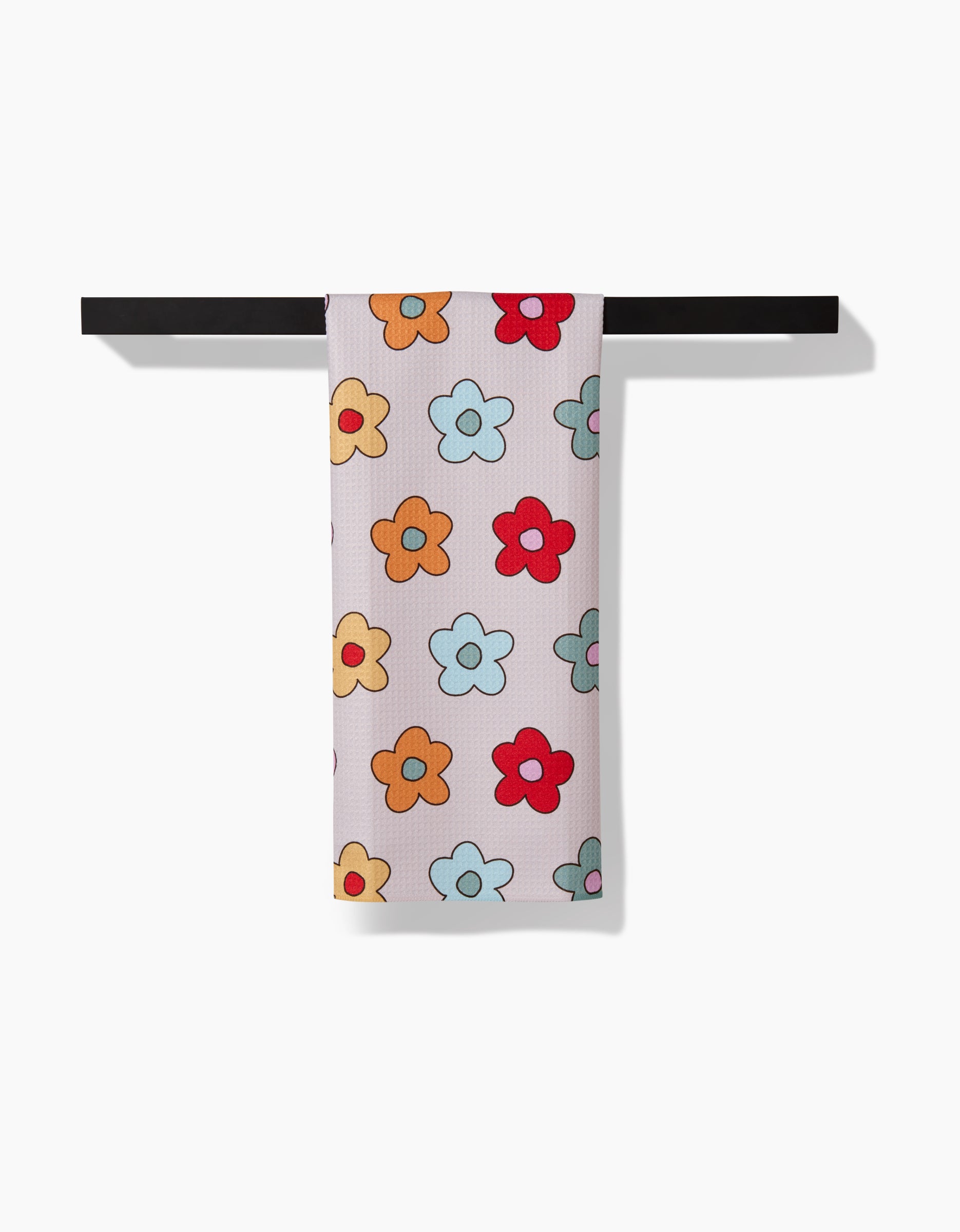 A Purely Kaylie Buttercream Blossoms tea towel, featuring vibrant cartoon florals in red, orange, yellow, and blue on white ultra-absorbent Geoweave™ fabric, hangs neatly over a black bar against a plain background.