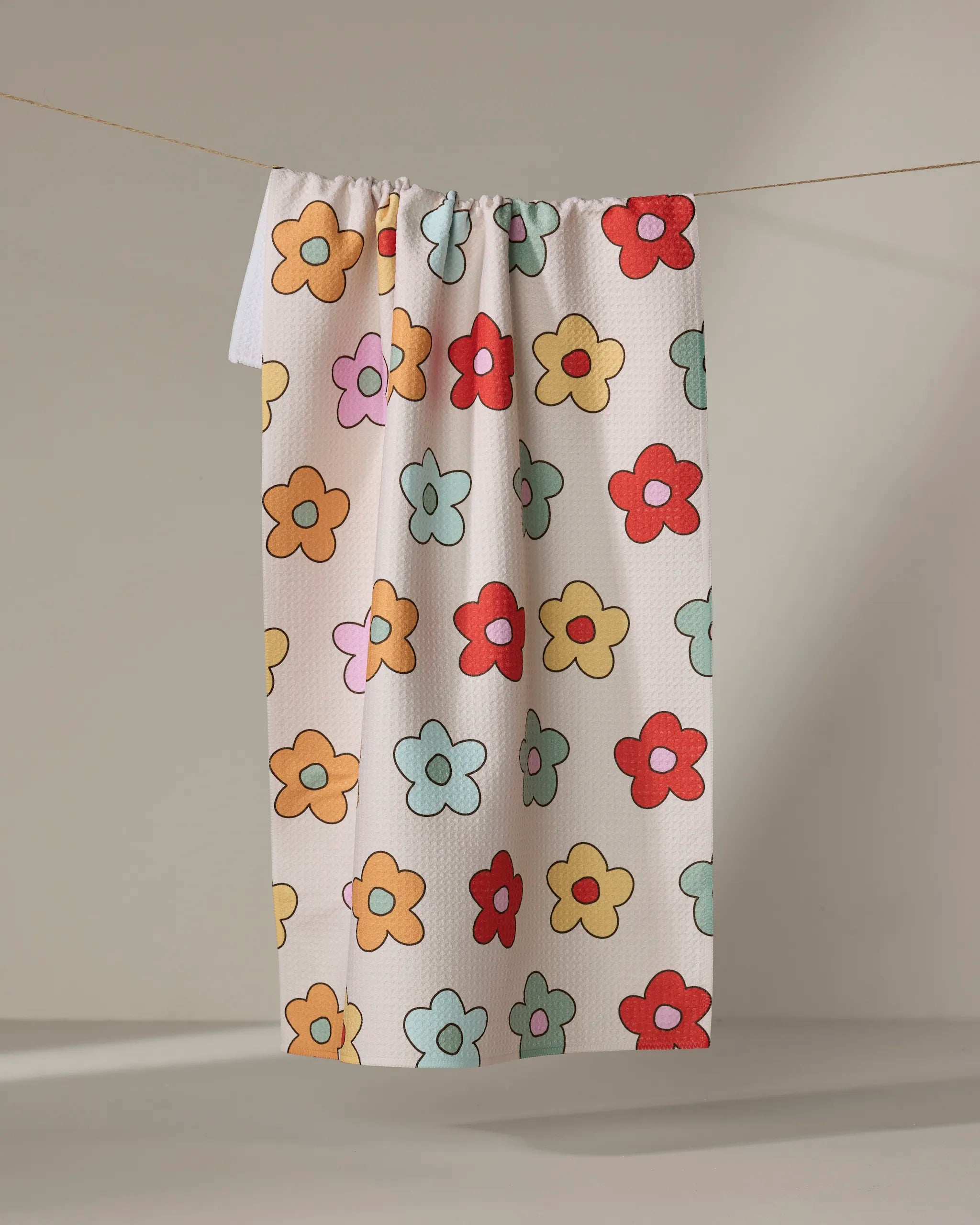 The Purely Kaylie "Buttercream Blossoms" kitchen towel, made from recycled materials, showcases a lively pattern of red, yellow, orange, pink, and light blue cartoon flowers hanging on a thin line against a plain background.