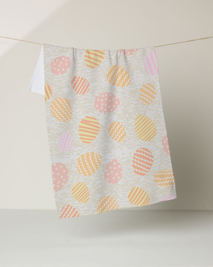 The Geometry Buttercream Easter tea towel, featuring a pink, orange, and yellow pastel egg pattern, hangs on a clothesline against a plain light background, adding brightness with its ultra-absorbent design.