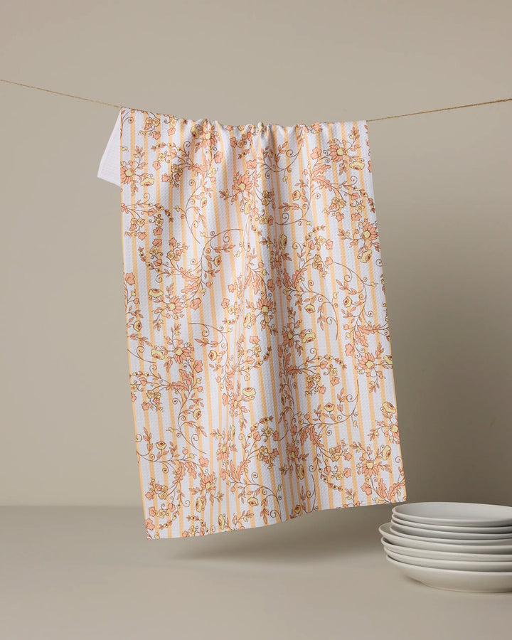 The Geometry Buttercup Vine kitchen towel, featuring an absorbent floral design with orange and yellow vines, hangs on a string against a beige background. Nearby, a stack of white plates rests on the surface.