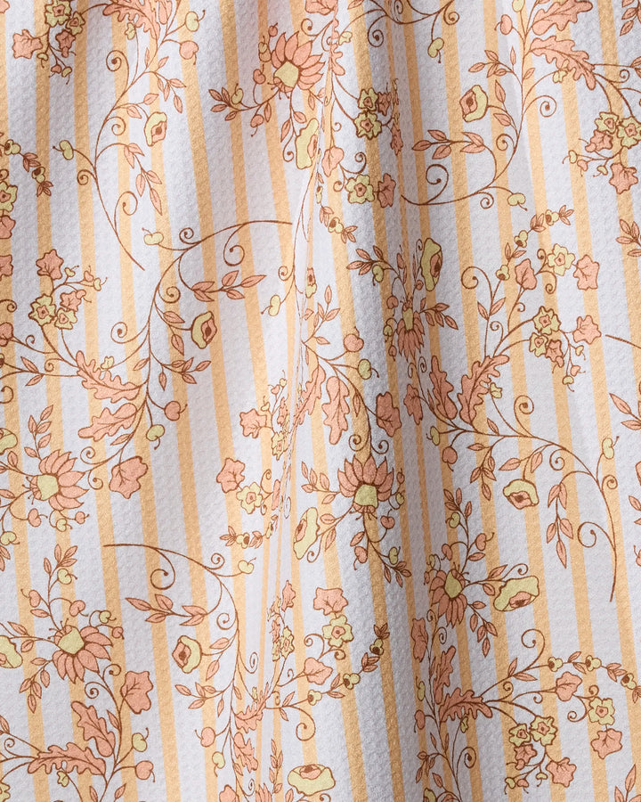 A close-up of Geometry’s Buttercup Vine tea towel shows vertical beige and white stripes with an orange and yellow floral pattern. The absorbent towel features a textured surface and is softly draped.