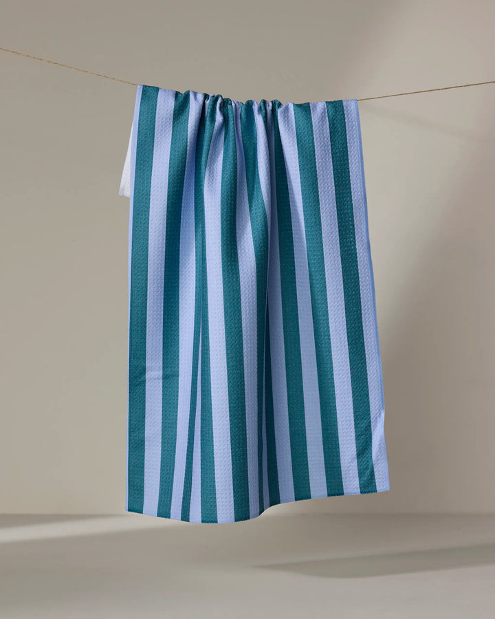 The Buy Guide’s Cabana Boy - Emerald French Blue tea towel, featuring blue and white vertical stripes, hangs on a clothesline against a plain beige background.
