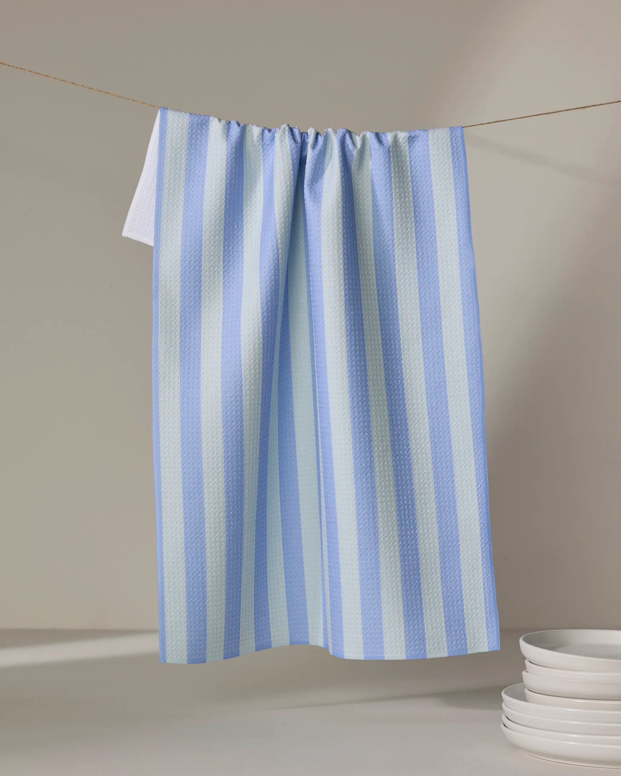 The Buy Guide's Cabana Boy - French Blue Mint tea towel, featuring blue and white stripes, hangs on a clothesline next to a stack of white plates on a light surface, set against a simple beige background.