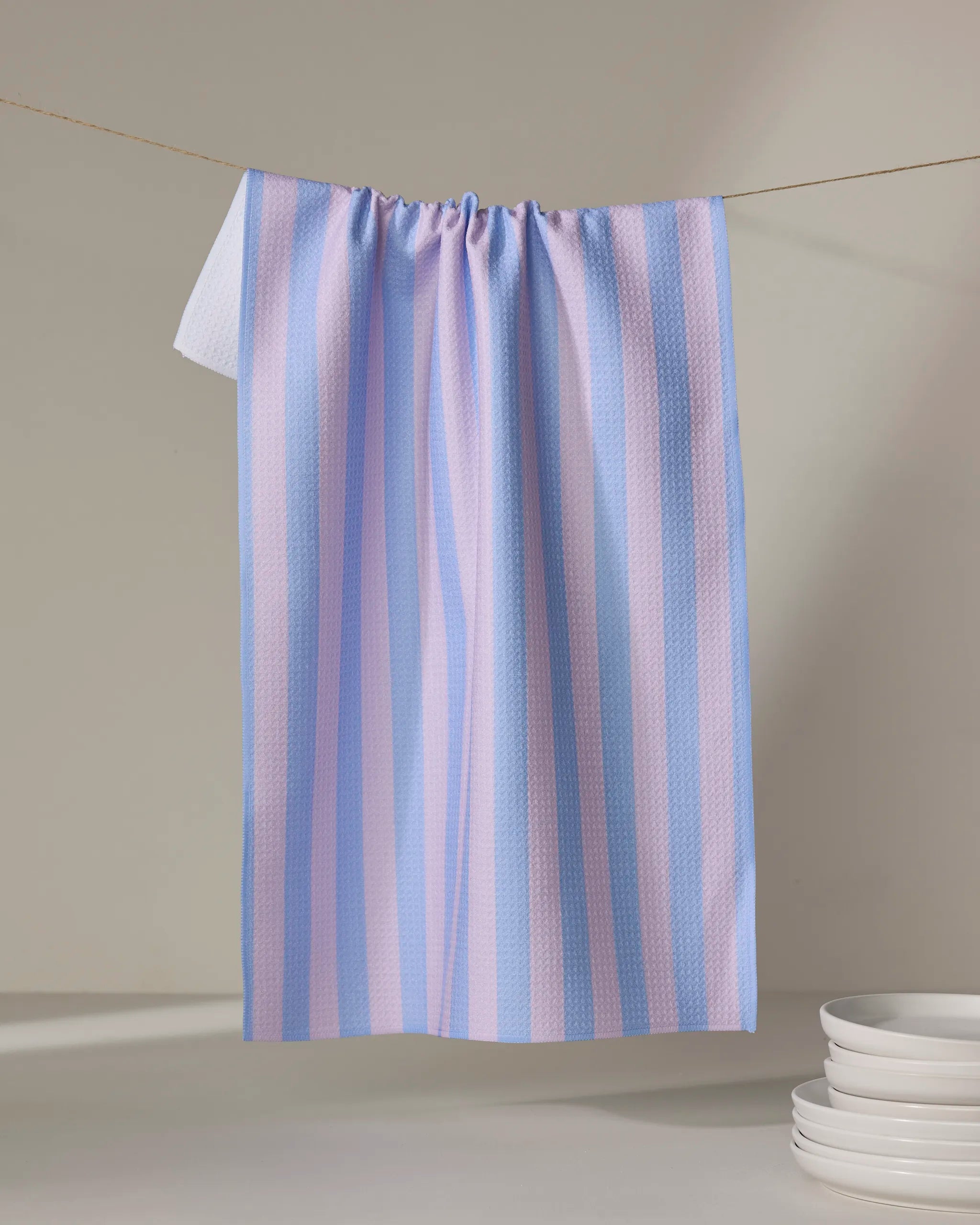 The Buy Guide’s Cabana Boy - Lilac French Blue tea towel, featuring lilac and French blue stripes and crafted from recycled materials, hangs on a line beside a stack of white plates in a softly lit, minimal scene.
