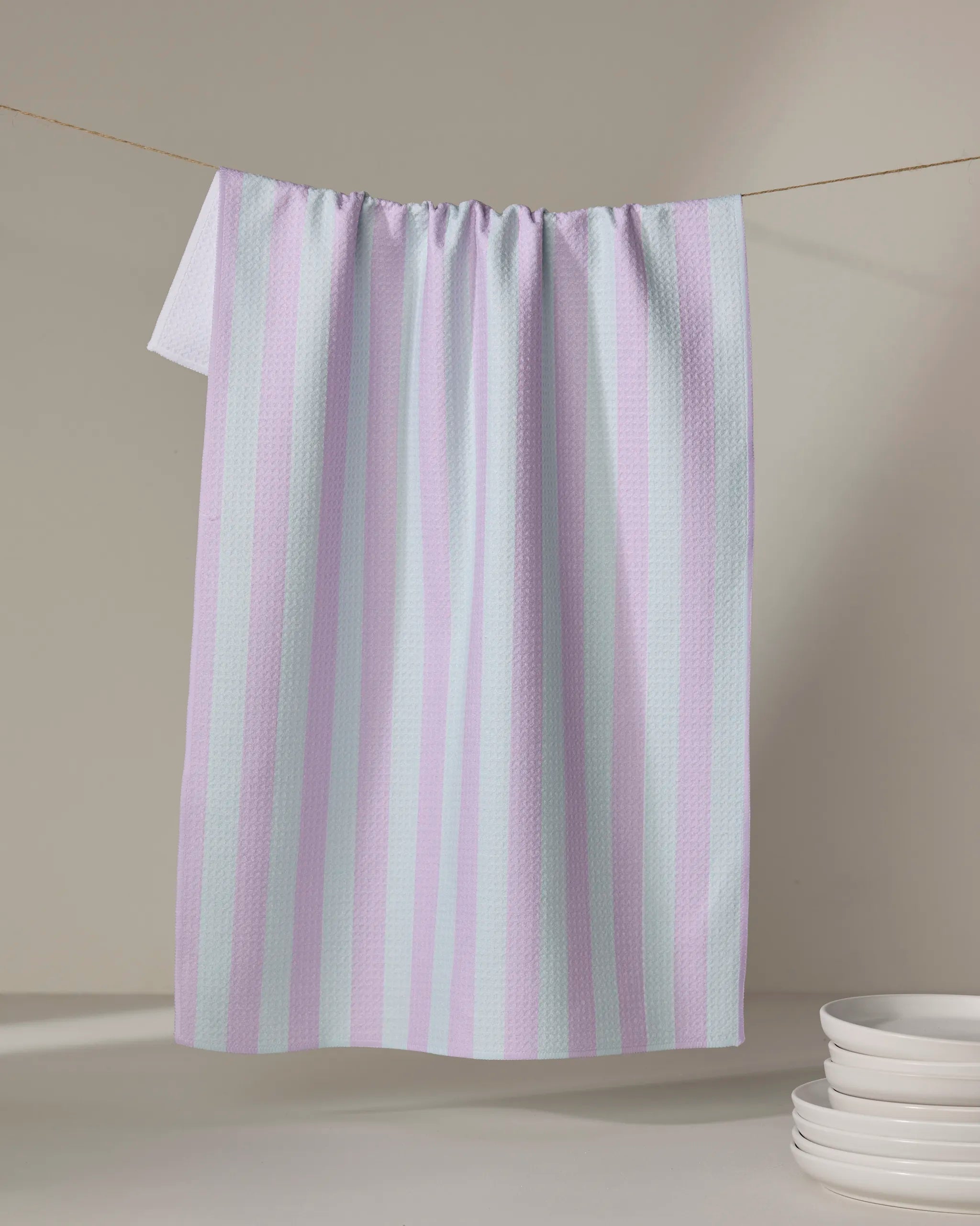 The Cabana Boy - Mint Lilac tea towel by The Buy Guide, featuring pastel pink and light blue stripes and made from recycled materials, hangs on a clothesline above stacked white ceramic bowls against a neutral backdrop.
