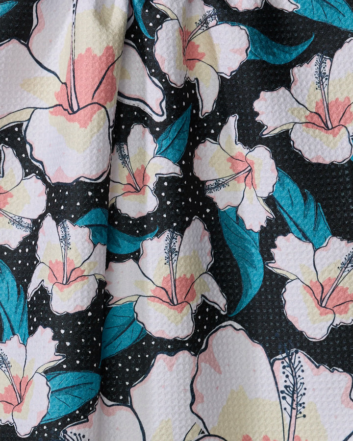 A close-up of the Cabo Cutie tea towel by Julianne Haness, made from recycled materials, featuring white and pink flowers, teal leaves, and small dots on a black background with a slightly textured look.