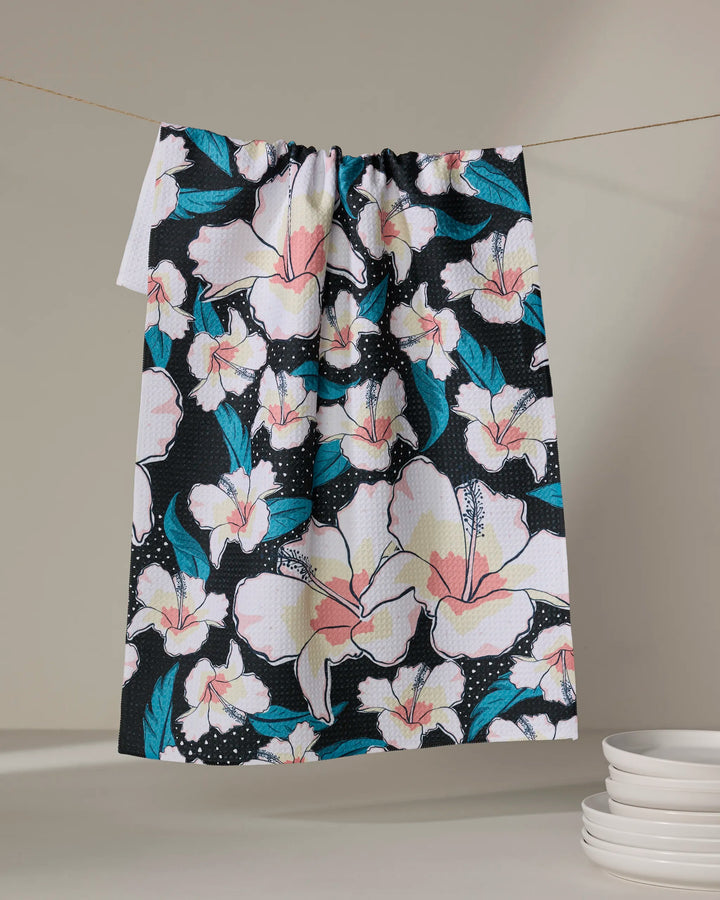 The Cabo Cutie tea towel by Julianne Haness features a vibrant pink, white, and blue floral pattern on black. Ultra-absorbent for everyday use, it hangs above white plates against a neutral backdrop.