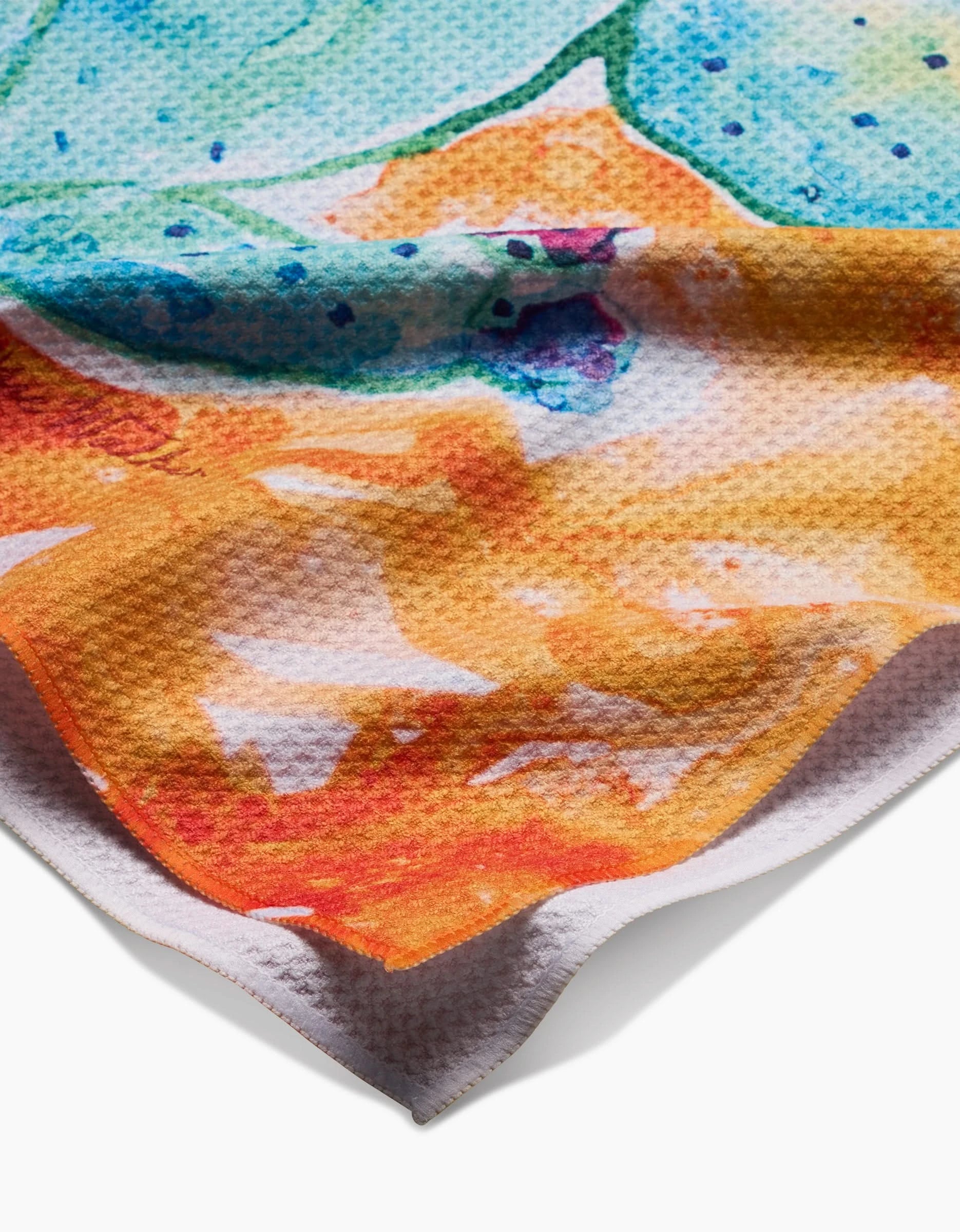A close-up of a slightly wrinkled Geoweave tea towel by Skye Walker, featuring Cacti—a vibrant abstract watercolor design in orange, yellow, blue, and green tones.