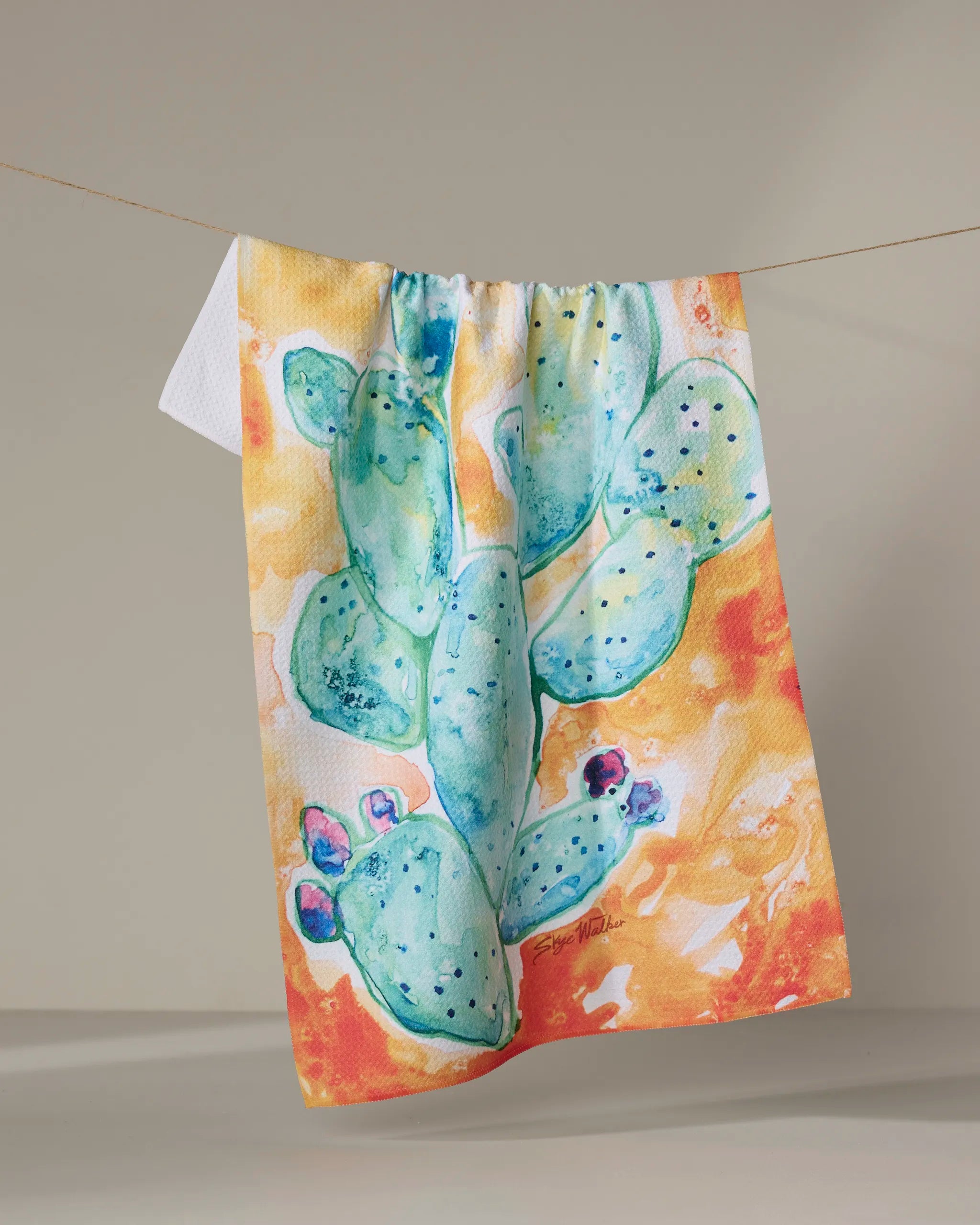 The Skye Walker "Cacti" tea towel showcases a quick-drying design with a vibrant watercolor green cactus and purple flowers on a beige background.
