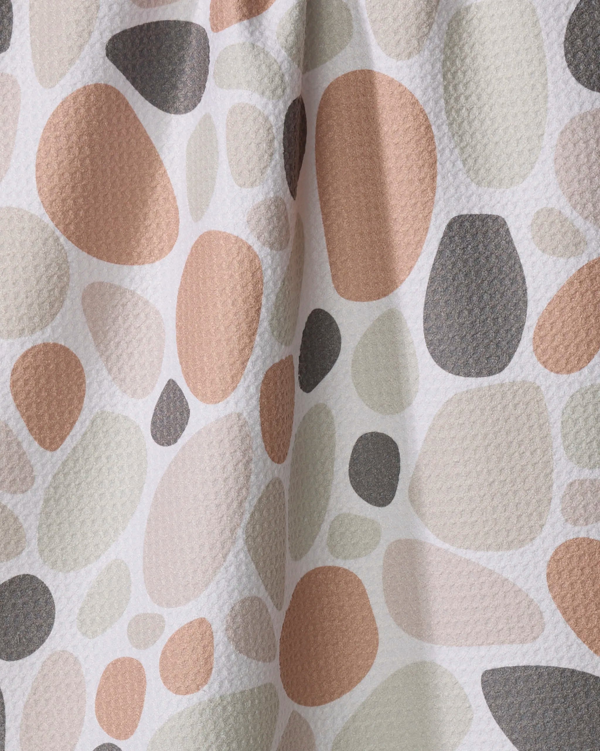 A close-up of textured fabric on the Rosa Kichline "Cactus Pieces" tea towel, featuring an abstract pebble pattern in muted beige, peach, taupe, and gray. Light and shadow ripple across its quick-drying material.