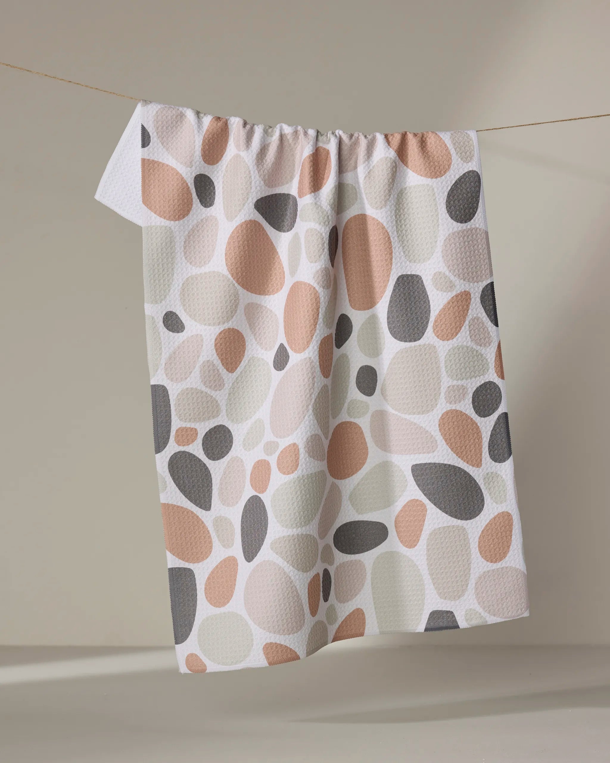 The "Cactus Pieces" tea towel by Rosa Kichline, featuring irregular pebble-shaped spots in beige, terracotta, gray, and white, hangs on a string against a neutral background.