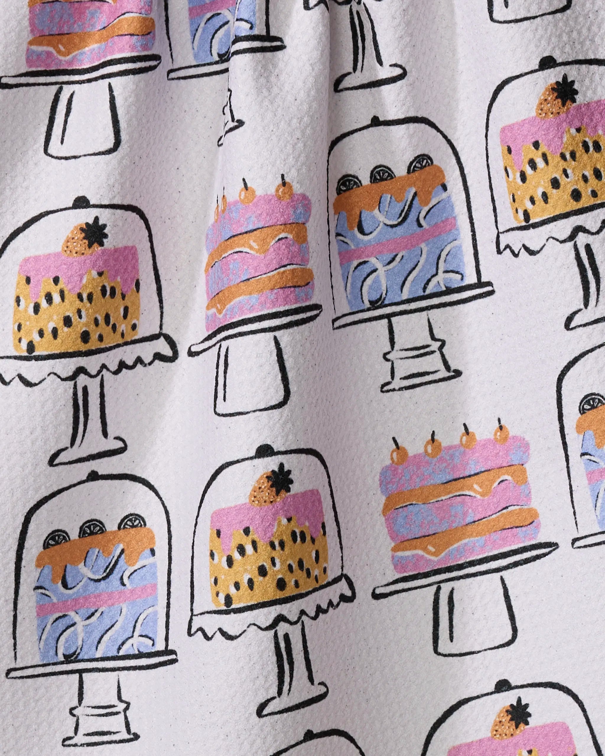 The "Cakes In The Window" tea towel by Megan Roy features colorful, hand-drawn cakes with pink, orange, purple, and yellow layers—decorated with fruit or dots—on cake stands against a light background.