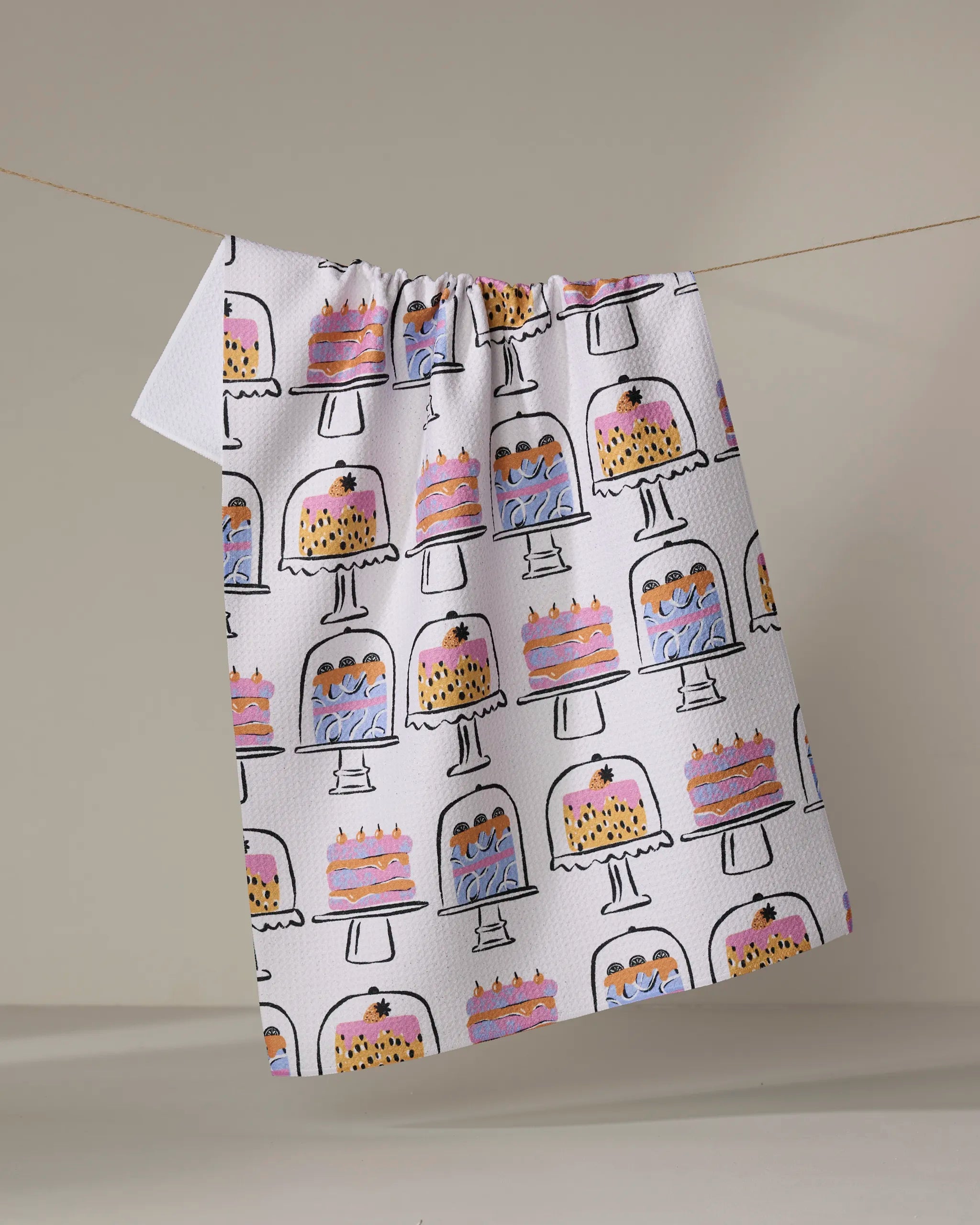 The "Cakes In The Window" tea towel by Megan Roy features vibrant cake illustrations on stands, crafted from recycled materials and displayed hanging against a plain background.