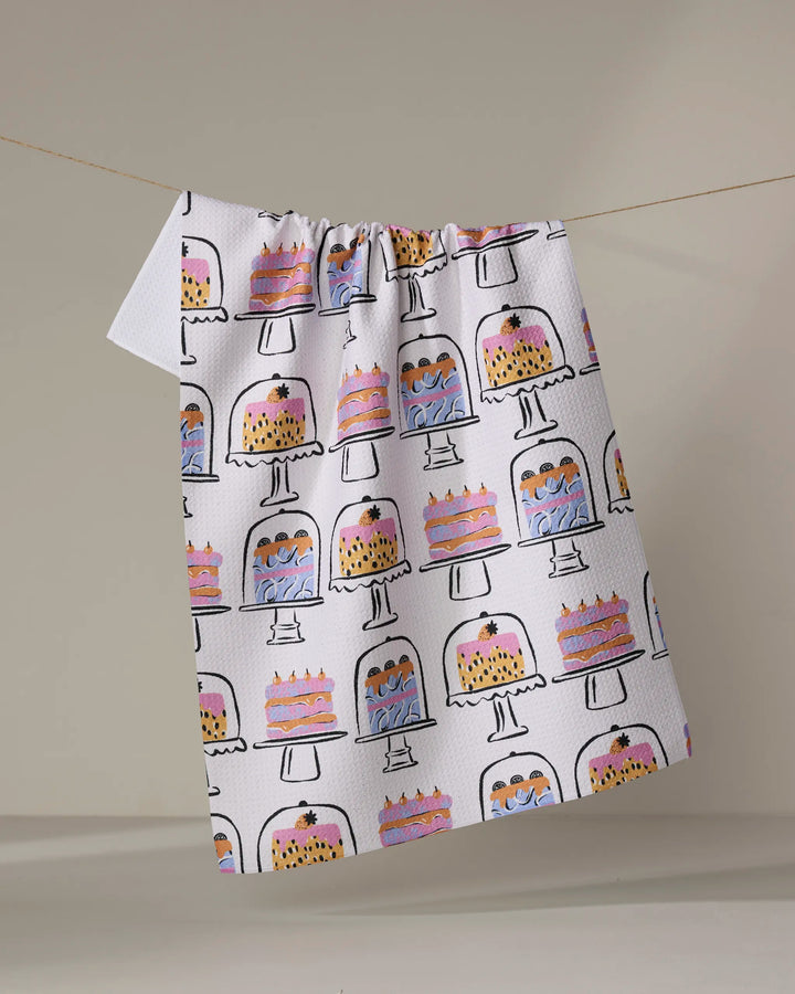 The "Cakes In The Window" tea towel by Megan Roy features vibrant cake illustrations on stands, crafted from recycled materials and displayed hanging against a plain background.
