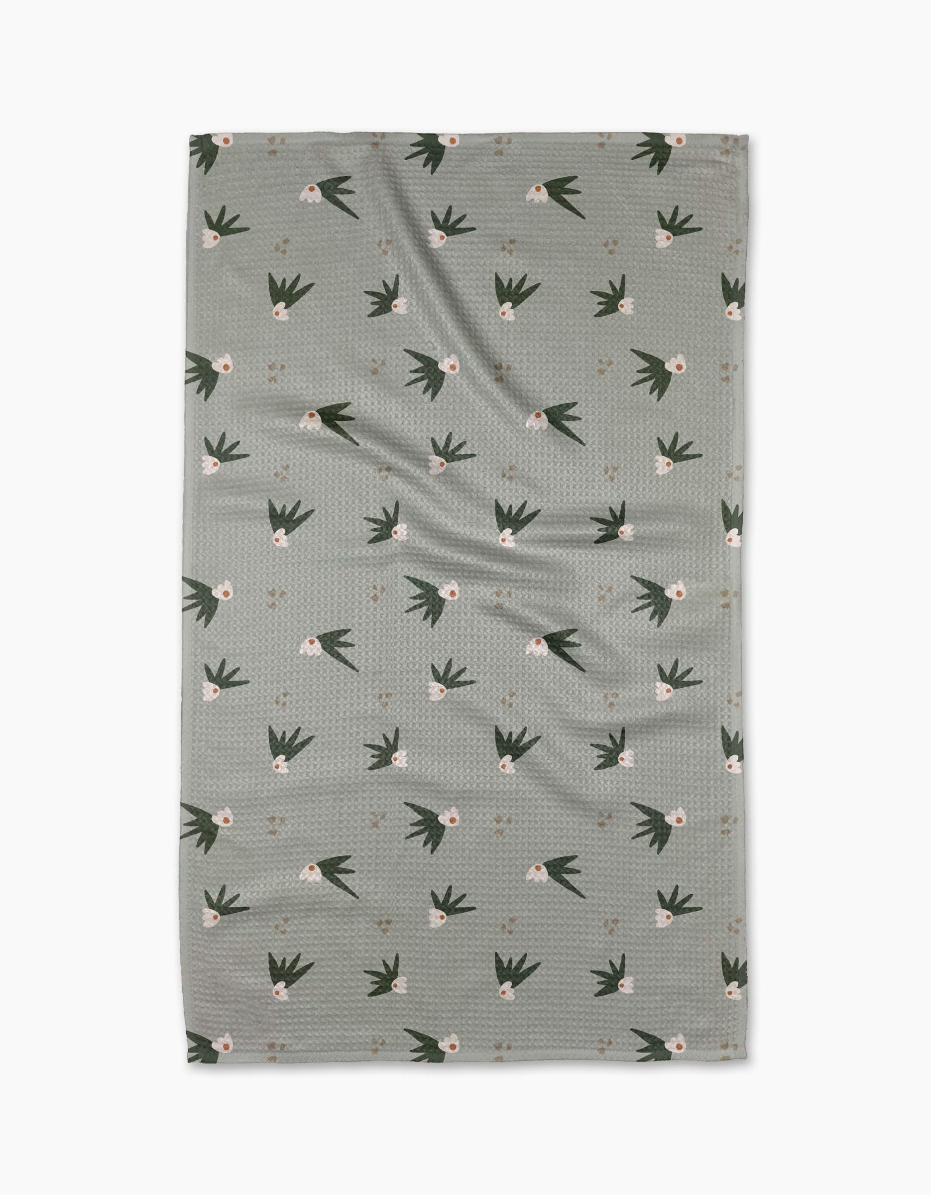 The Cali Sun Agave tea towel by Amberleigh Adoff features a grayish-green rectangular design with a subtle texture, scattered white flowers, and dark green leaves, expertly crafted from recycled materials.