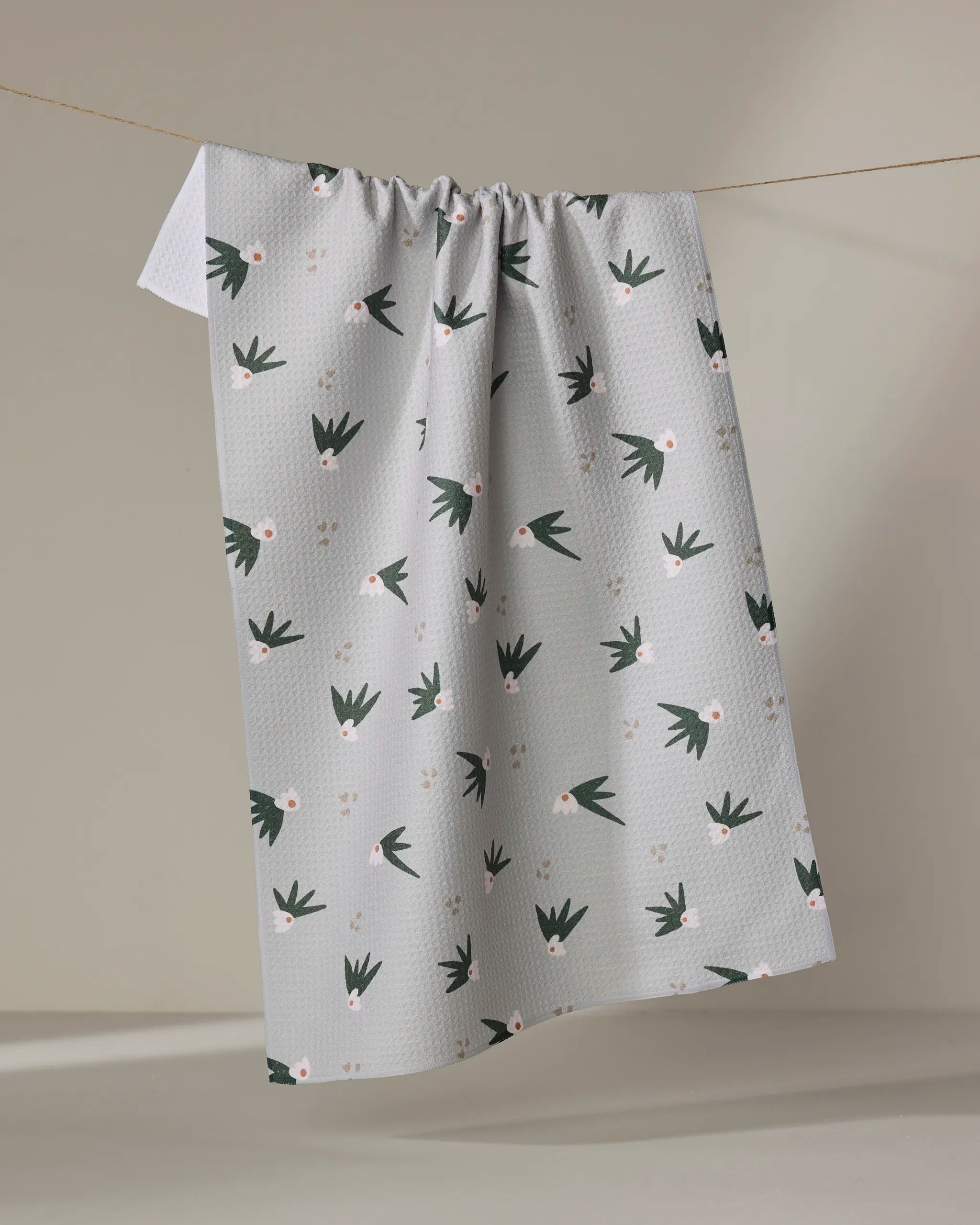 The Cali Sun Agave kitchen towel by Amberleigh Adoff, featuring a light gray grid pattern with green leafy shapes and small pink flowers, hangs on a string against a neutral background.