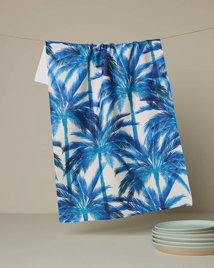 The Caligo tea towel by Geometry, crafted from recycled materials and featuring a blue palm tree design, hangs on a thin string against a neutral background, with white plates stacked in the lower right corner.