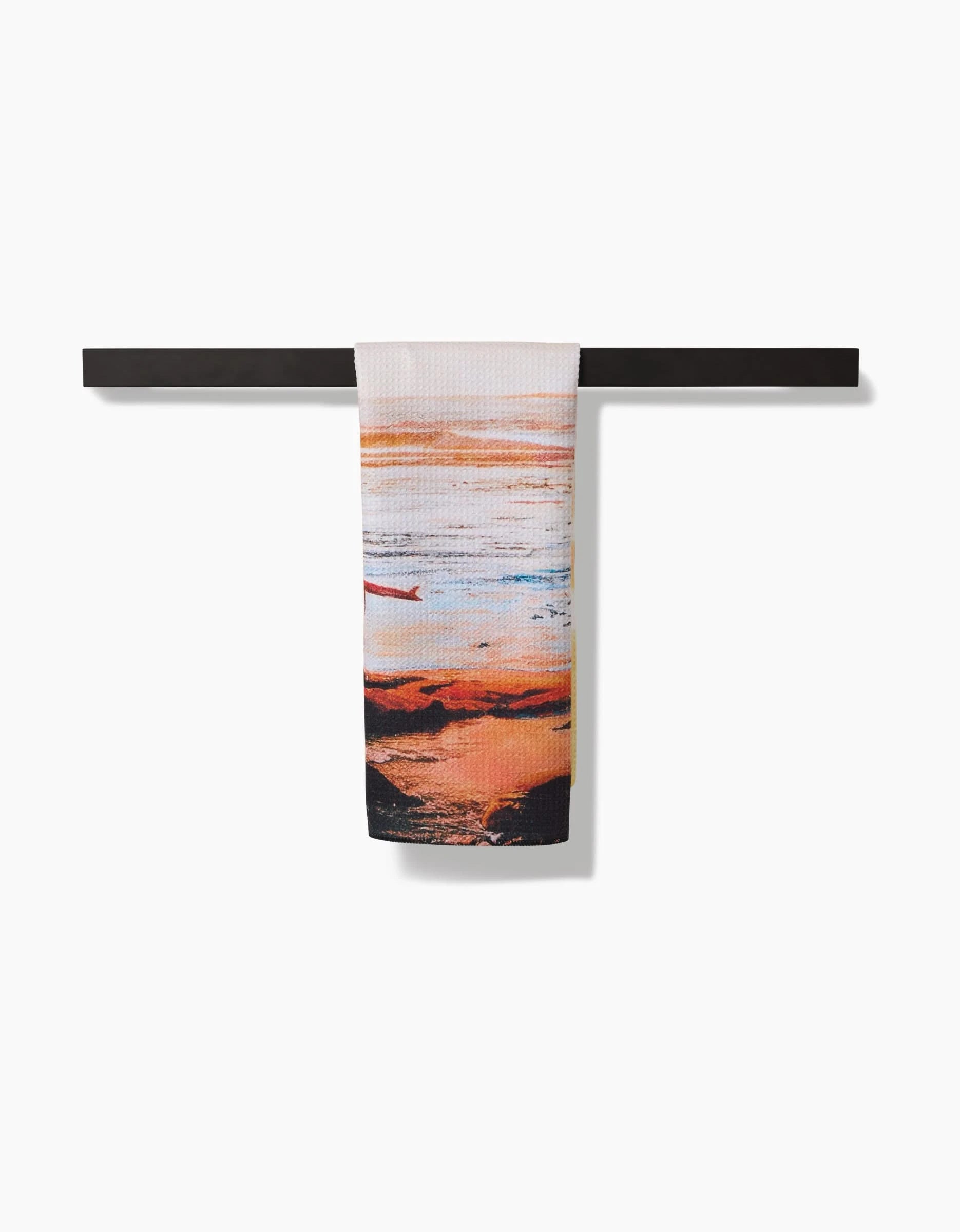 The Call Of The Wild tea towel by Stefanie Bales, made from soft Geoweave fabric, displays a sunset beach scene with a small boat and hangs over a sleek black bar against a white background.
