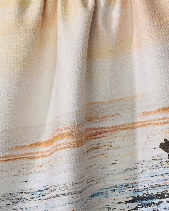A close-up of the Stefanie Bales "Call Of The Wild" tea towel shows fabric gathered at the top, featuring an ultra-absorbent material and an ocean sunset design in orange, yellow, and blue over water.