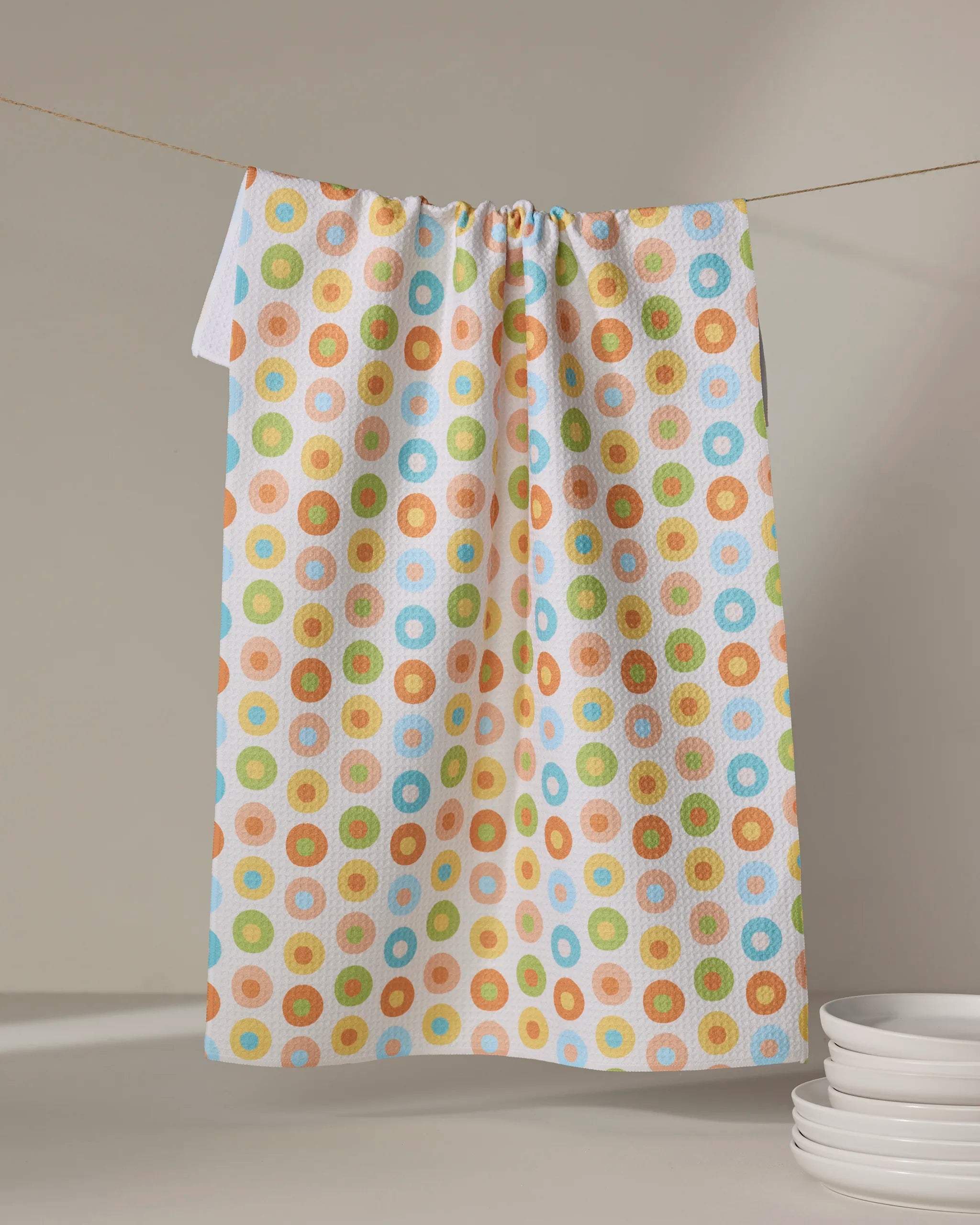 The Mindfulnice Candy Dots tea towel, made from recycled materials and featuring colorful polka dots, hangs on a clothesline with a stack of white plates in the lower right corner.