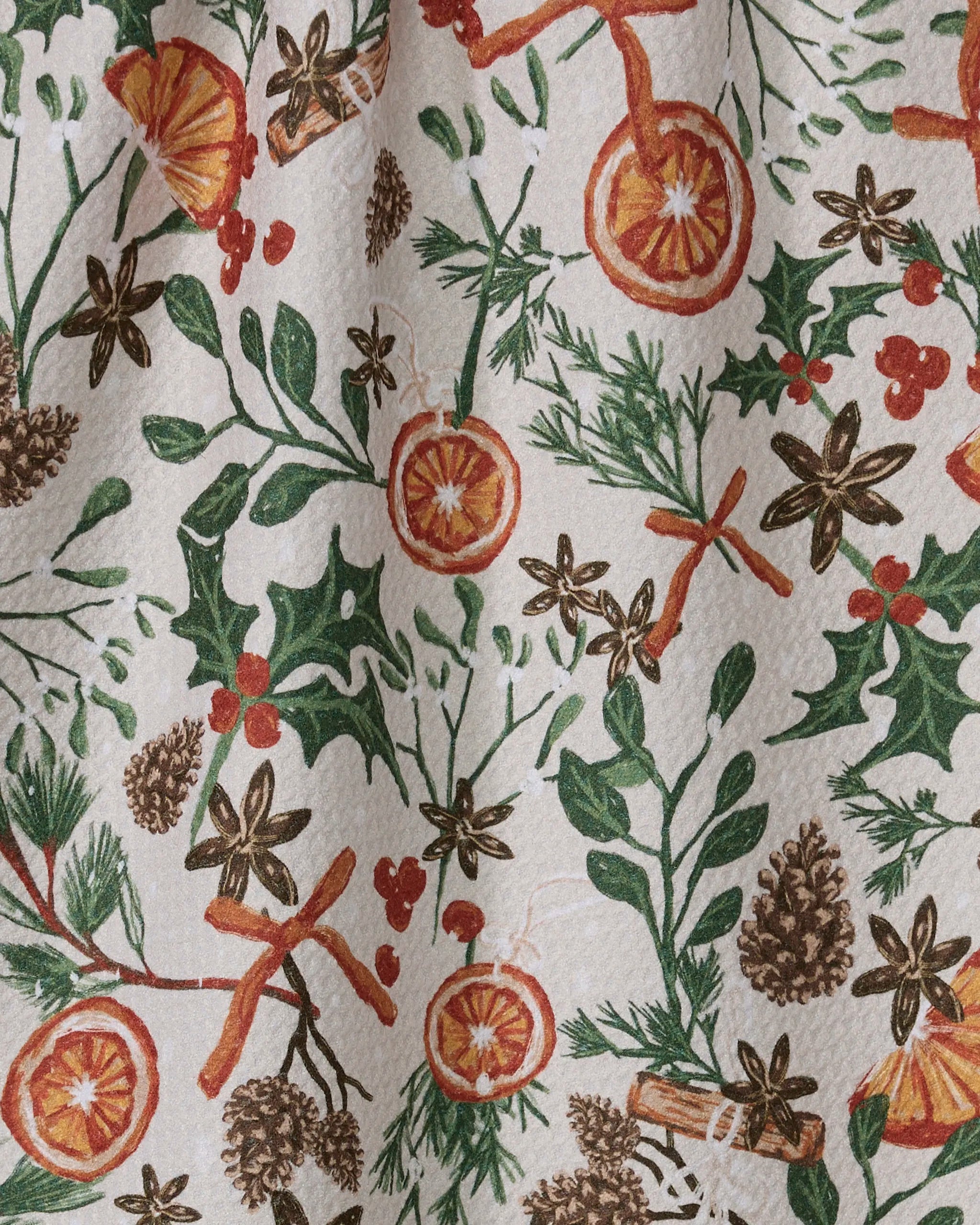The Geometry Canela kitchen towel features a festive pattern of pinecones, holly leaves, cinnamon sticks, star anise, orange slices, berries, and green foliage on a textured white background.