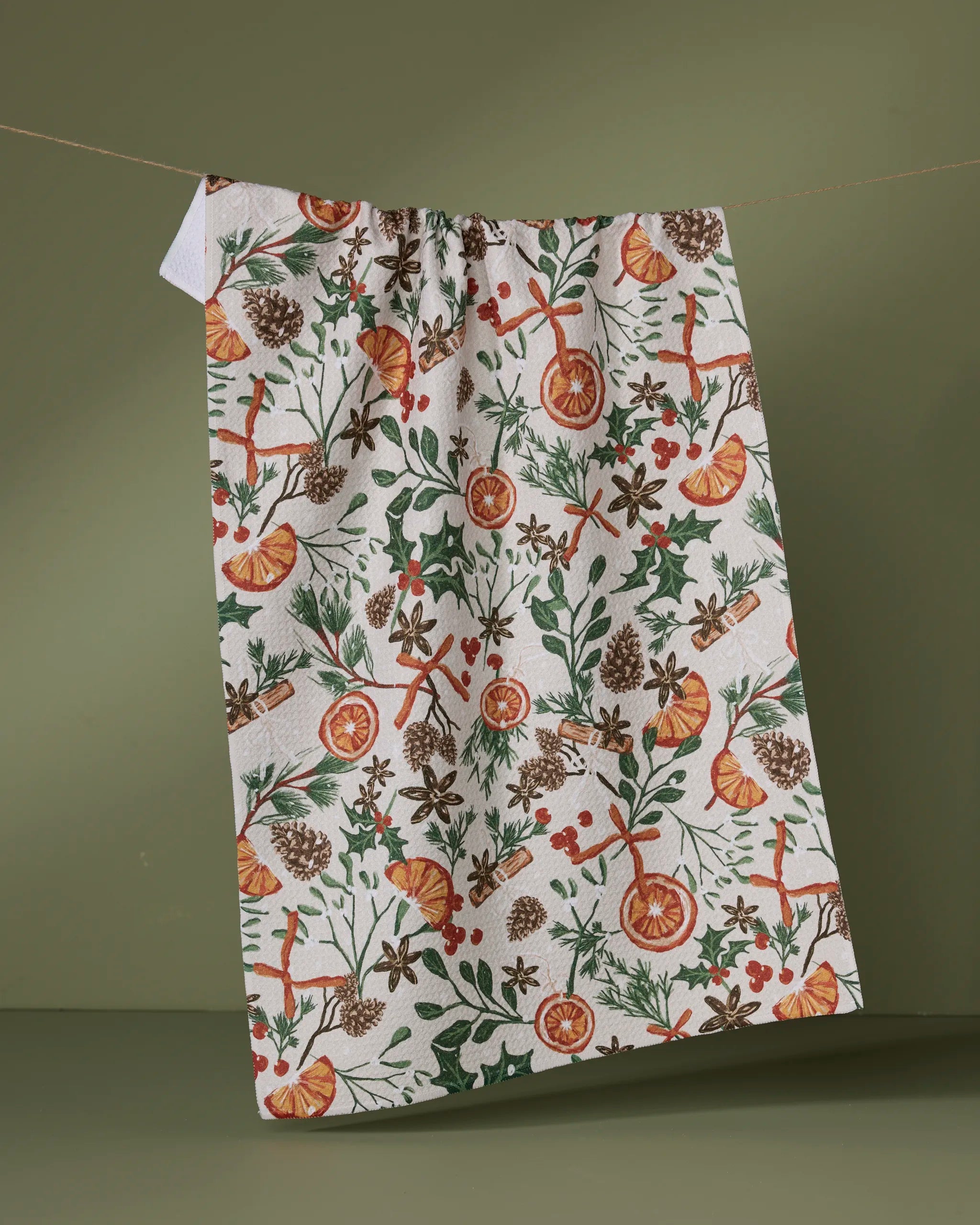 The Geometry Canela kitchen towel, made from recycled materials and featuring a festive botanical print with orange slices, pinecones, cinnamon sticks, berries, and green foliage, hangs on a string against an olive green background.