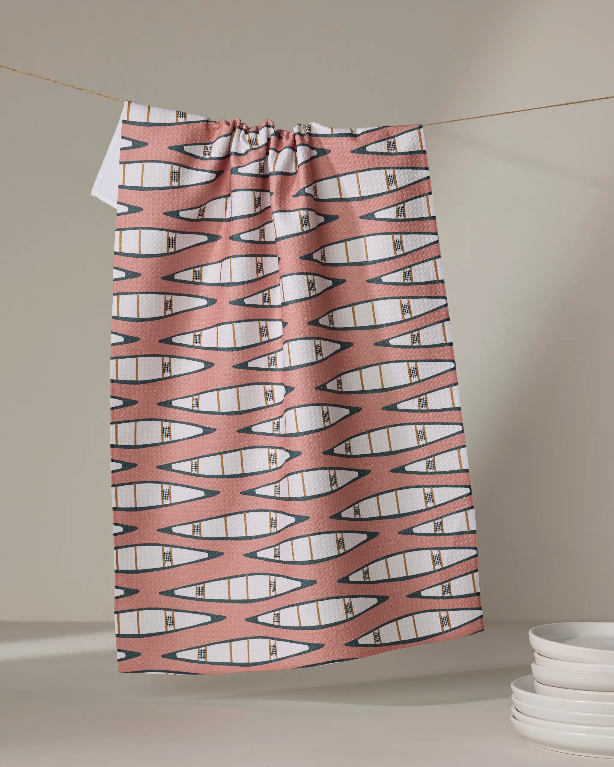 A "Canoes" tea towel by Coit Creative, featuring a pink and white geometric zigzag design and made from quick-drying recycled materials, hangs on a string near stacked white plates against a neutral backdrop.