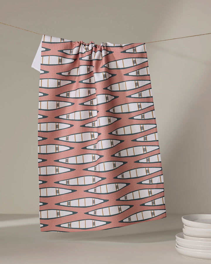 A "Canoes" tea towel by Coit Creative, featuring a pink and white geometric zigzag design and made from quick-drying recycled materials, hangs on a string near stacked white plates against a neutral backdrop.