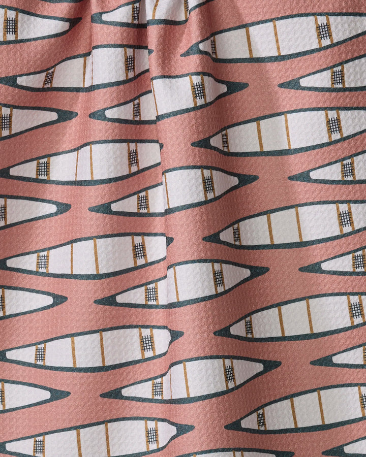 The Coit Creative "Canoes" tea towel features a repeating pattern of white canoes outlined in black and tan on a muted pink background. Made from recycled materials, the fabric includes subtle folds and gentle shadows.