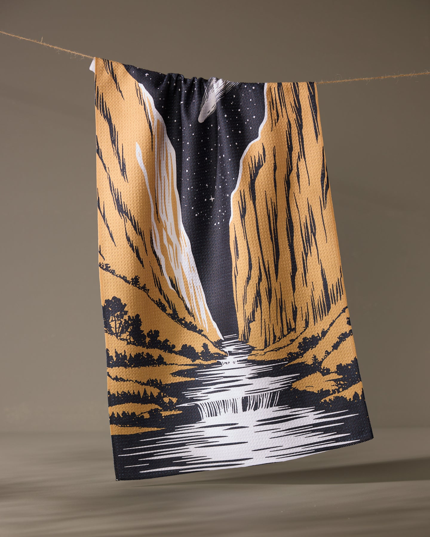 The Canyonfall kitchen towel by Geometry features an illustrated river winding between golden cliffs under a starry night sky, hanging from a clothesline against a plain background.