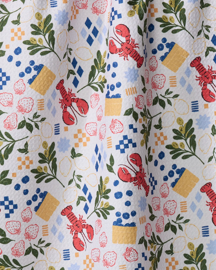The Capri kitchen towel by Charlie Rowan Designs features a playful pattern of red lobsters, strawberries, lemons, green herbs, blue and yellow checkered shapes, and small blue dots on recycled fabric.