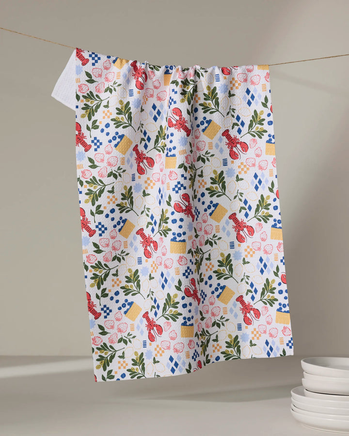The Capri dish towel by Charlie Rowan Designs, featuring a vibrant pattern of lobsters, flowers, sponge cakes, and leaves, hangs on a clothesline above stacked white plates.