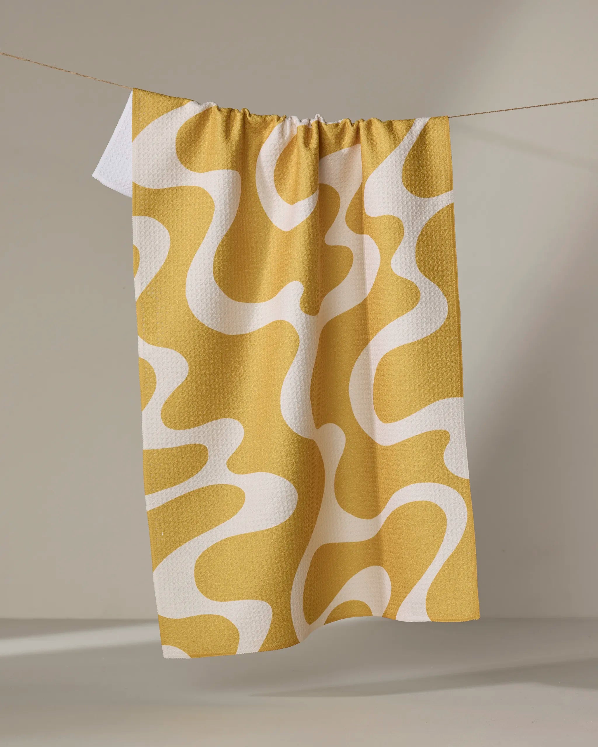The Gerdadzy Caramel Swirl tea towel, in yellow and white with a wavy abstract design, hangs on a string against a neutral background. Made from recycled materials, its textured waffle surface ensures ultra-absorbency.