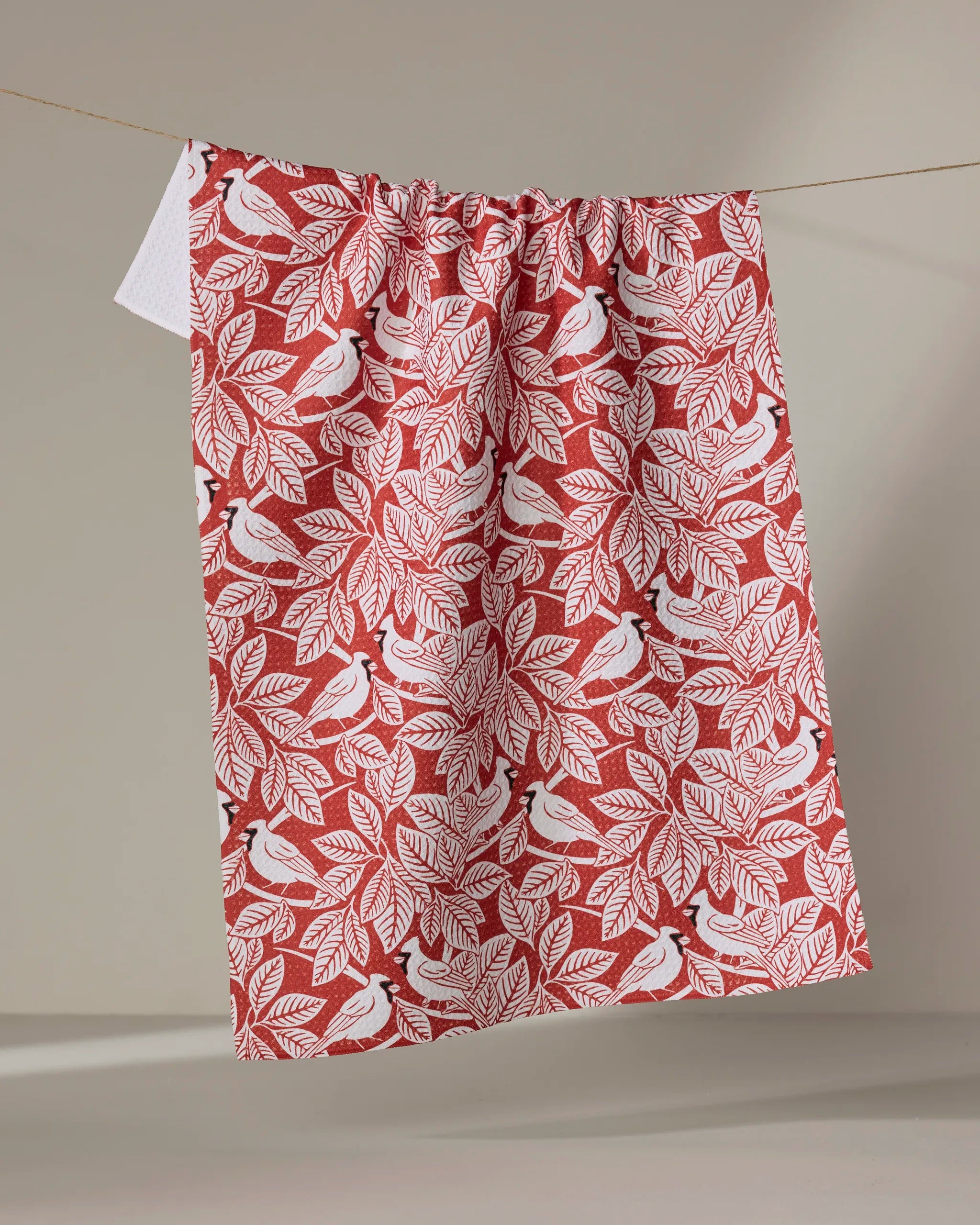 The Cardinal Canopy tea towel by Sarah Daugherty features birds and leaves in red and white, hangs on a clothesline, and is made from quick-drying recycled materials.