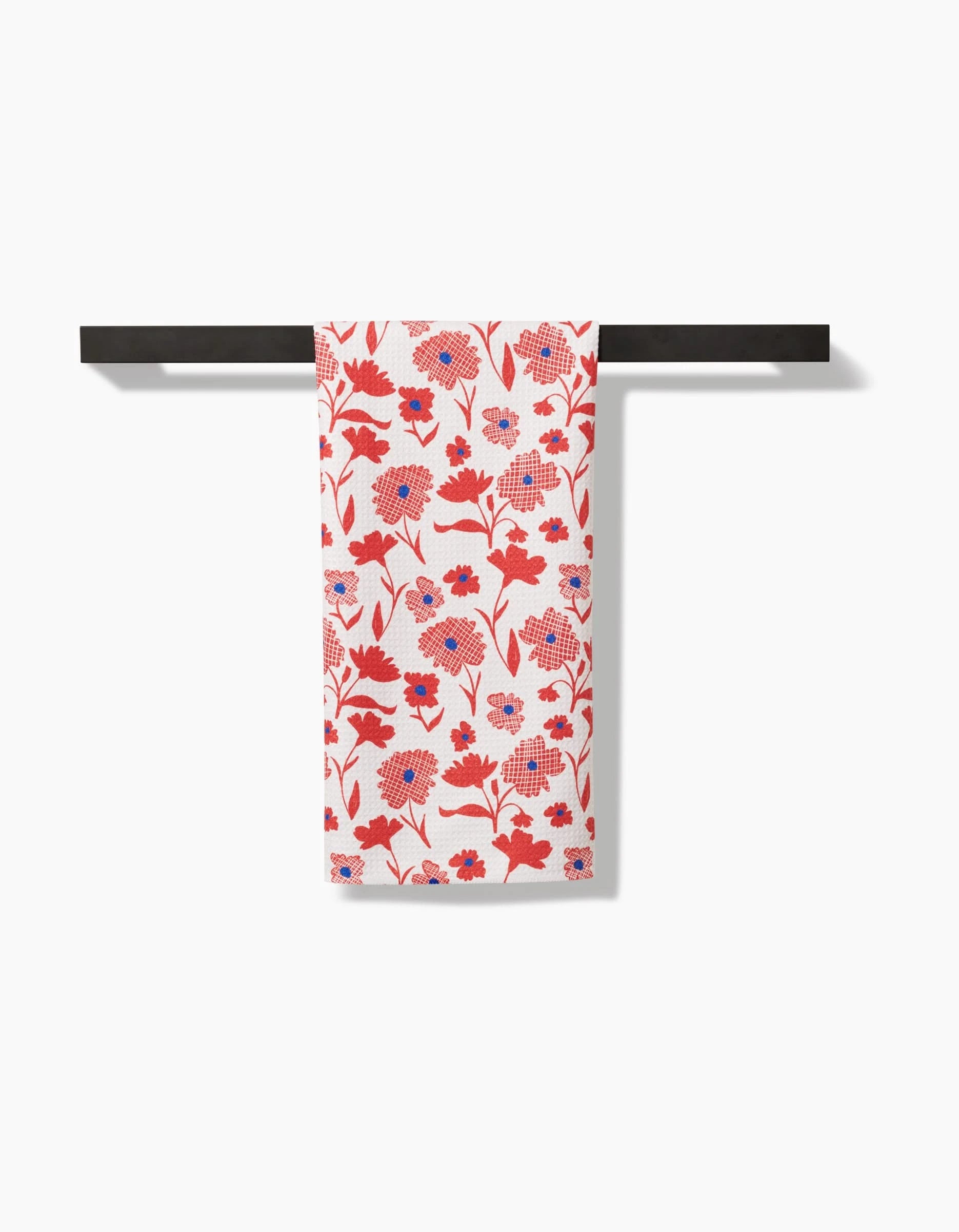 The Carmine tea towel by Danelys Sidron, crafted from premium Geoweave fabric for superior absorbency, features a red and blue floral design on white and hangs neatly on a black towel rack against a plain white background.