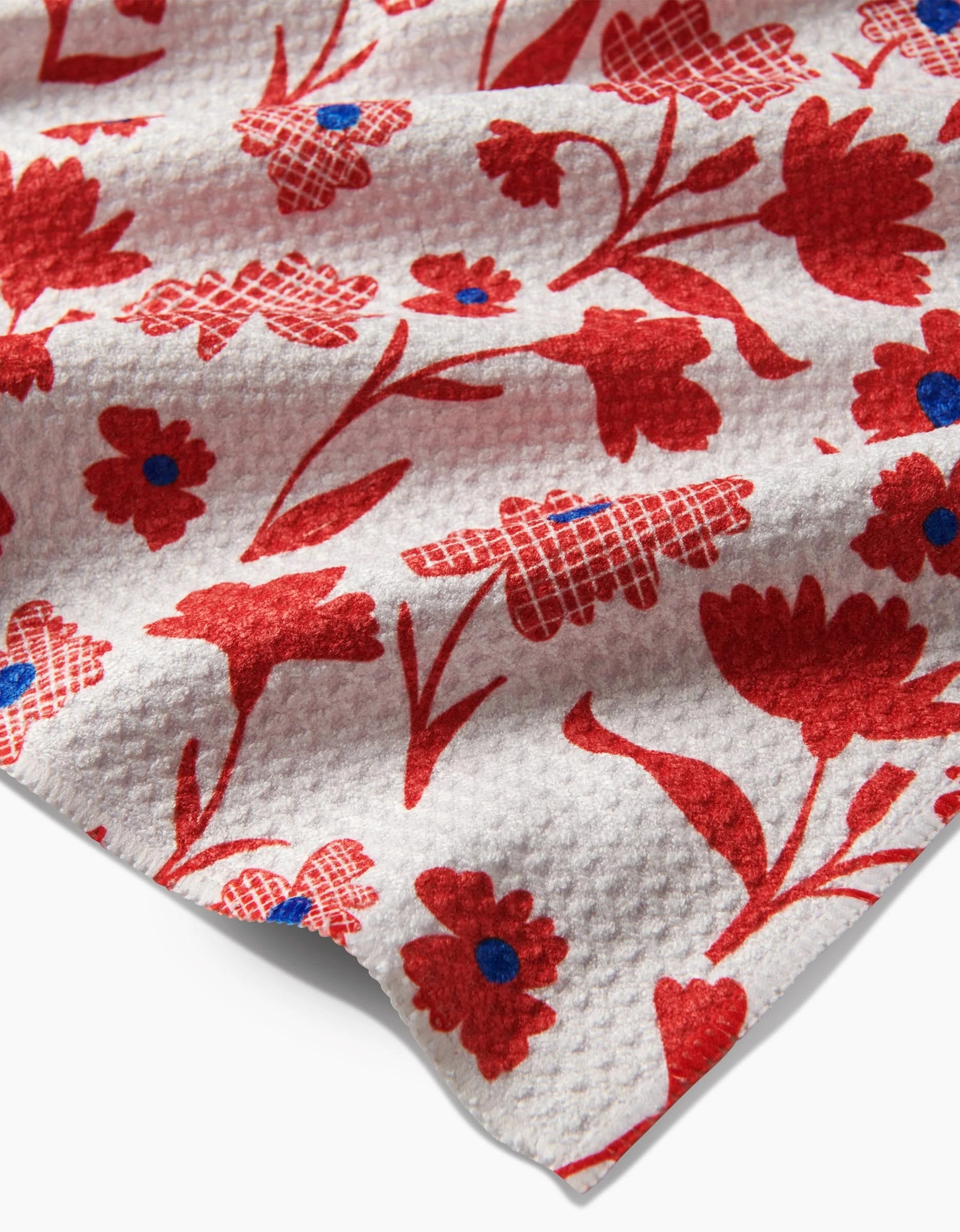 The Carmine tea towel by Danelys Sidron is made of white Geoweave fabric, featuring a textured pattern of red flowers with blue centers and checkered petal designs in various floral shapes.