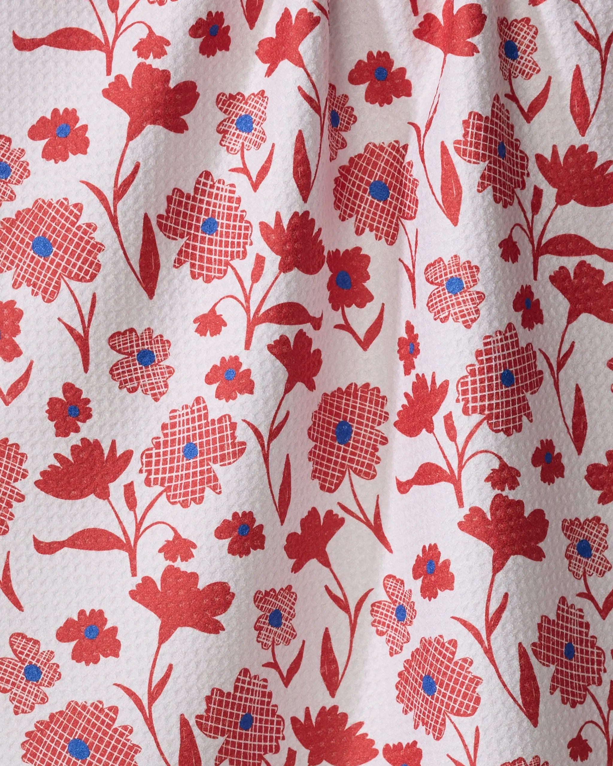 The Carmine kitchen towel by Danelys Sidron features an absorbent, textured white fabric adorned with various red flowers, blue centers, and checkered details in a vibrant floral pattern.