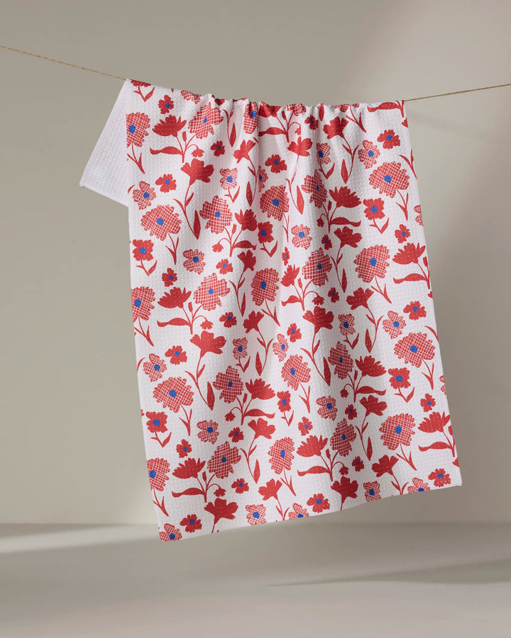 The Carmine kitchen towel by Danelys Sidron, featuring a red and blue floral pattern on white, hangs on a clothesline against a neutral background.