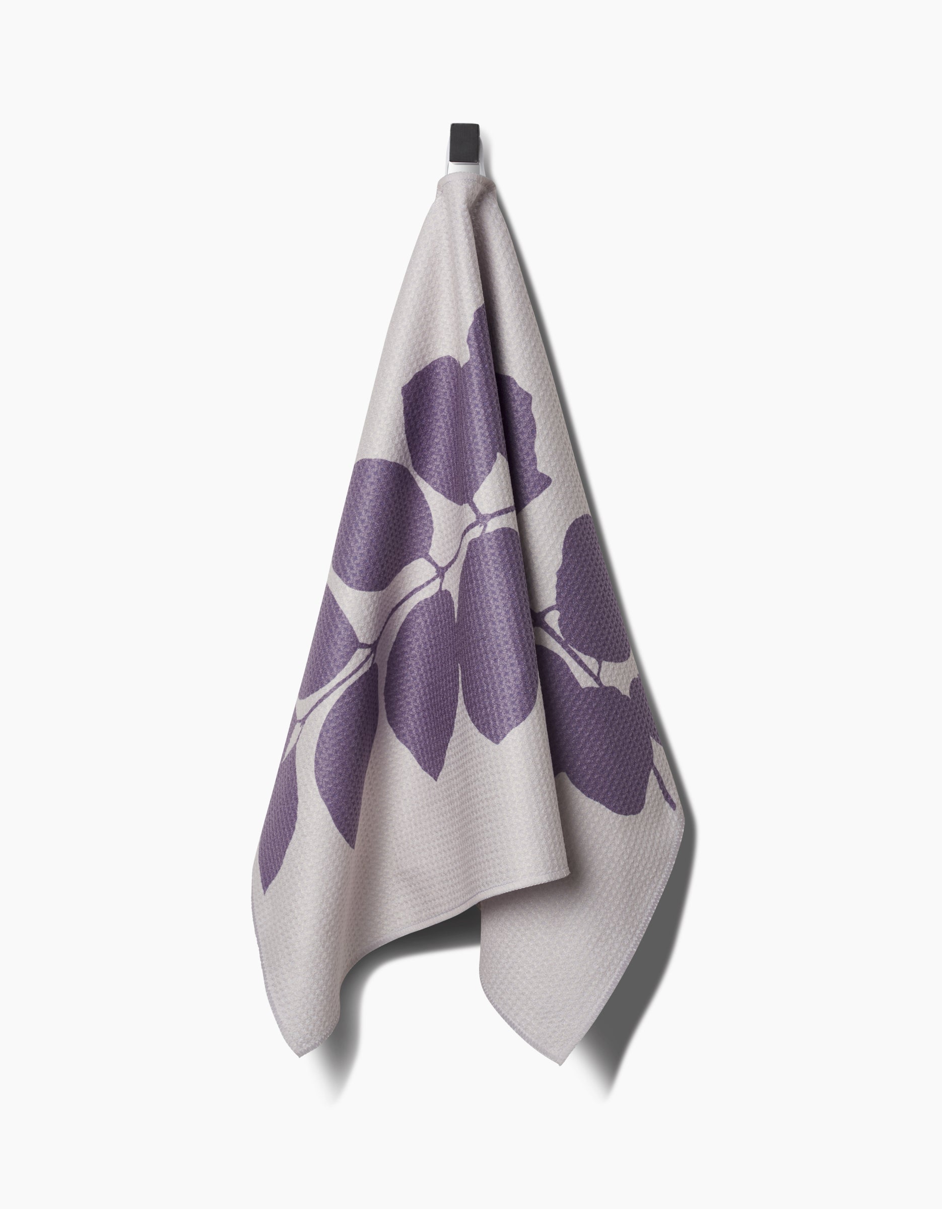 The Carob towel by PrintedPlant hangs on a hook, showcasing a bold purple leaf design on light gray. Made from ultra-absorbent Geoweave fabric, it offers both style and superior drying performance.