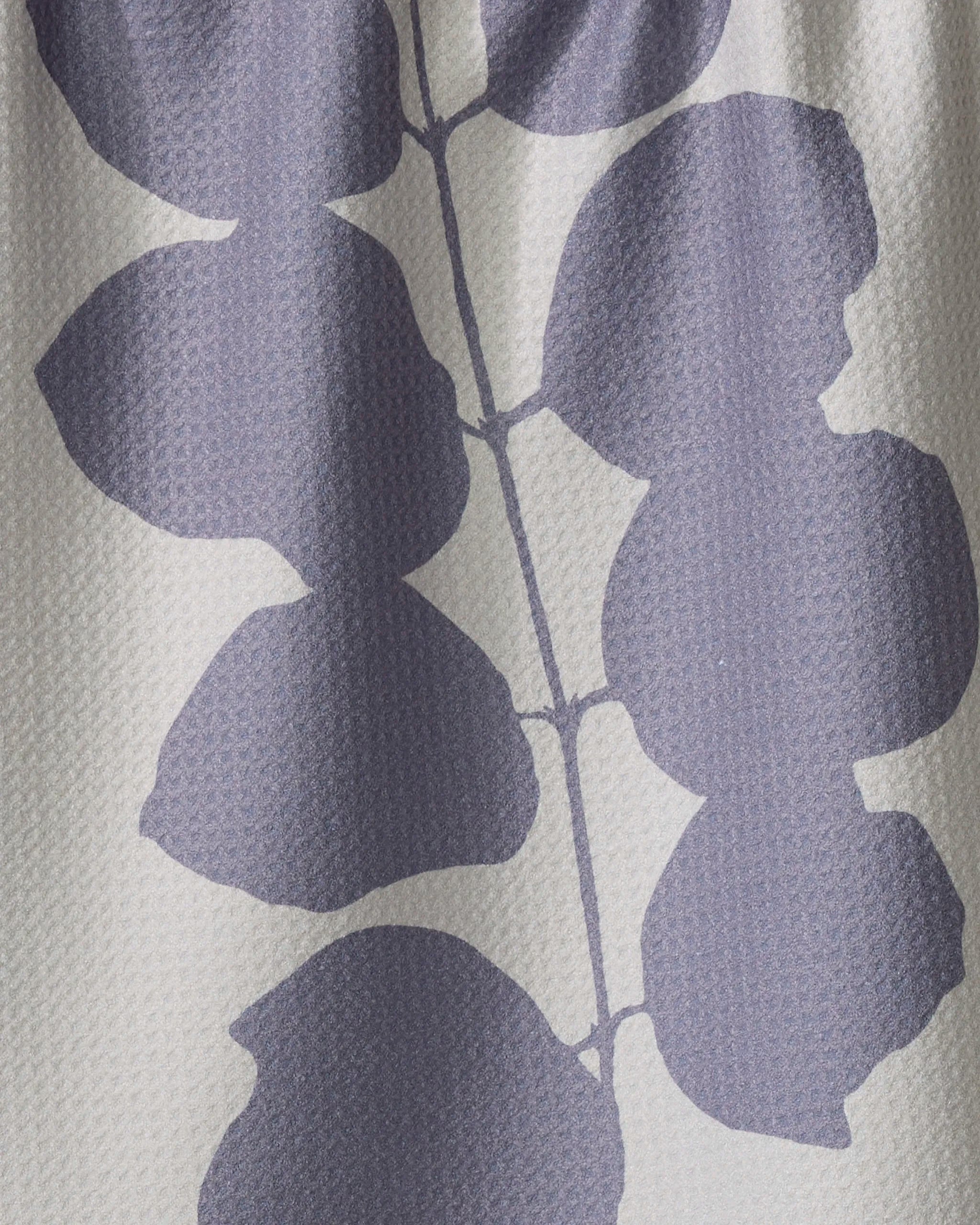 A close-up of PrintedPlant’s Carob recycled kitchen towel with a textured surface, showcasing large, abstract purple leaves and stems in a minimalist botanical pattern on a light beige background.